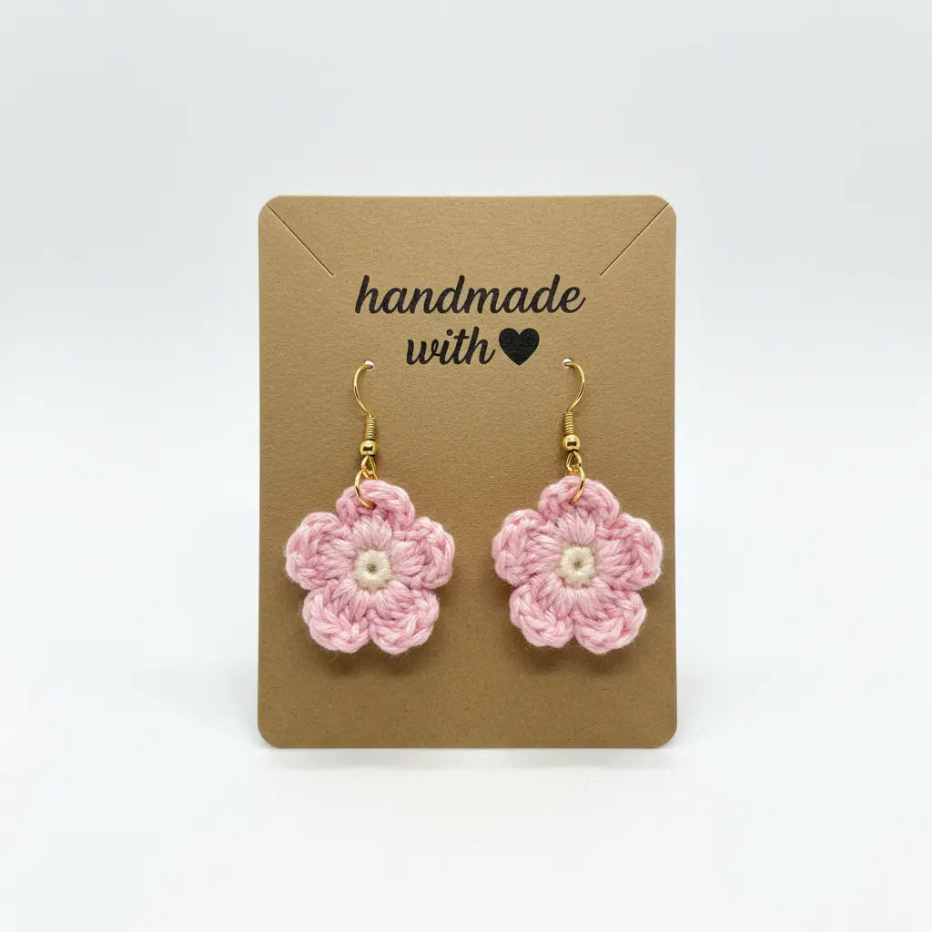 A pair of handmade pink crochet flower dangle earrings with gold hooks and white centers, displayed on a brown kraft card that reads 'handmade with' against a white background.