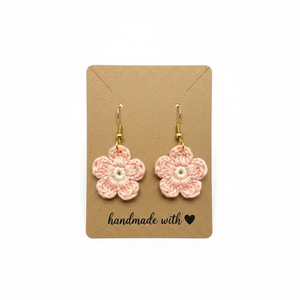 A pair of handmade crochet flower drop earrings in light pink and white, hanging from gold hooks on a brown kraft paper display card that reads 'handmade with heart'.