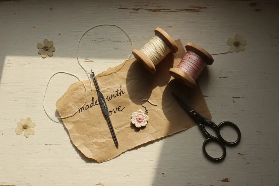 Behind the scenes at Loopelle Studio — crochet hook, yarn spools, scissors and a handmade micro crochet flower earring on kraft paper reading "made with love" on a rustic white wood surface