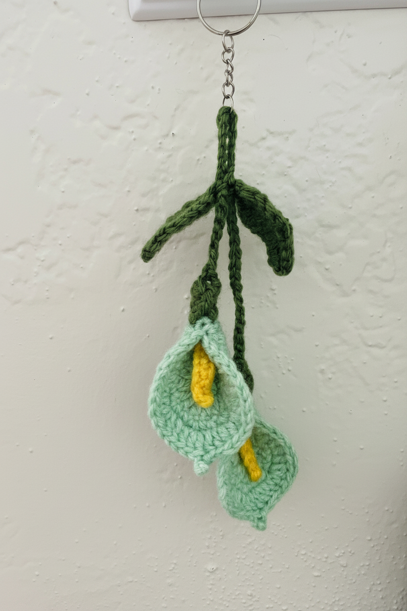 A crocheted hanging ornament shaped like calla lilies, with light green petals, yellow centers, and dark green stems, hanging from a metal ring against a textured off-white wall.