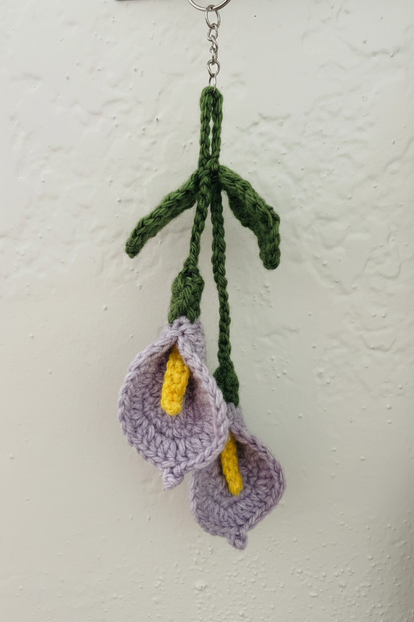 Hand-crocheted calla lily wall hanging ornament with three purple flowers, yellow centers, and green stems, suspended from a metal ring against a textured off-white wall.