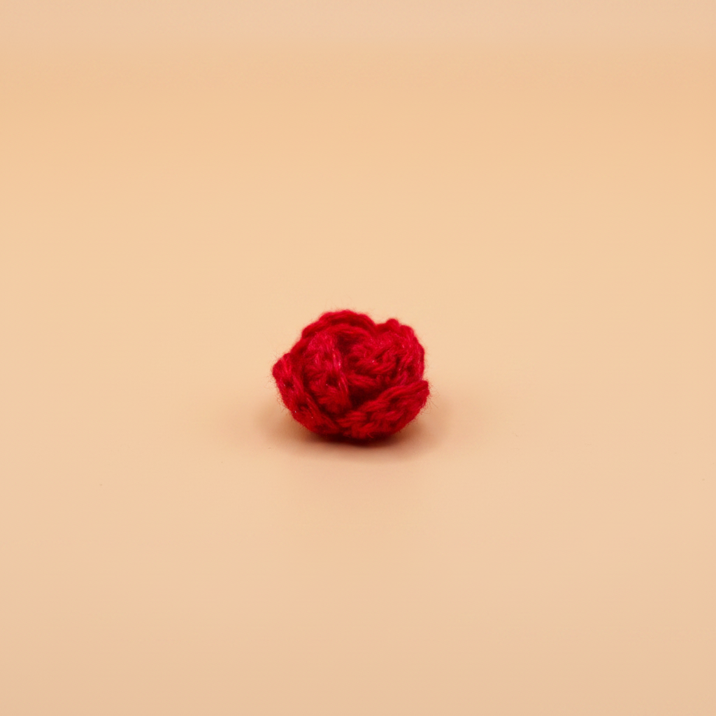 A small, textured red crochet or knitted rose-shaped accessory, possibly a hair scrunchie or decorative pom-pom, centered on a solid warm beige background.