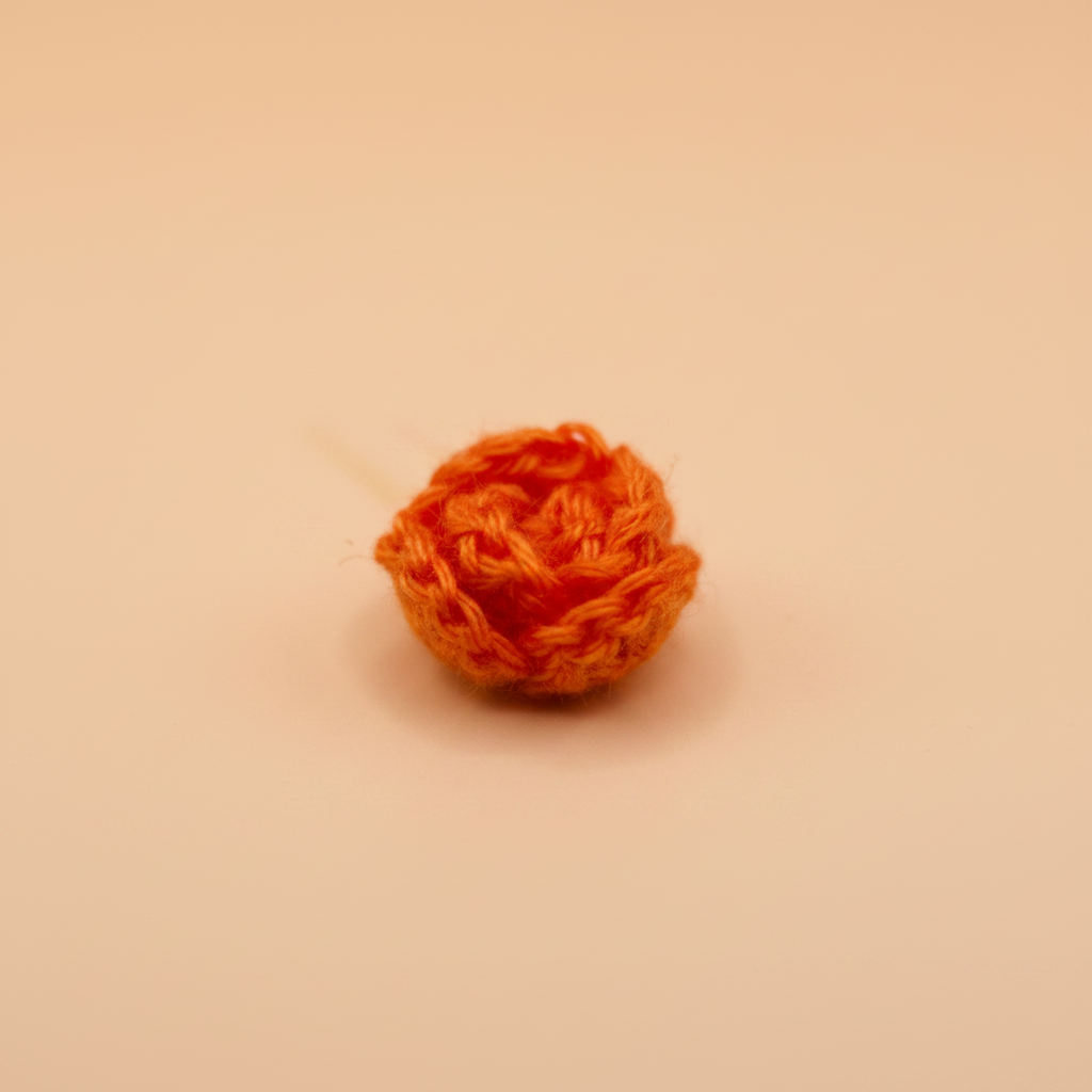 A small, hand-crocheted orange flower bud or rose-shaped decorative piece on a solid beige background.