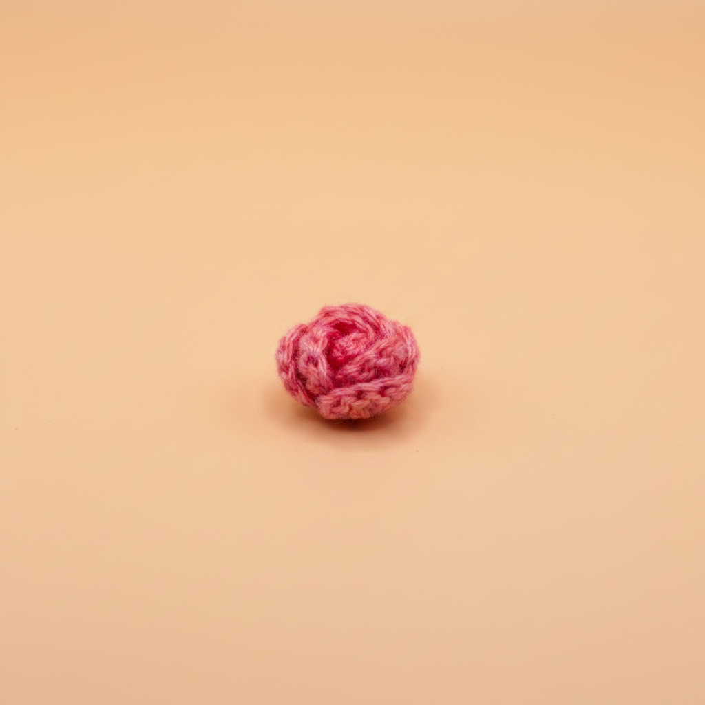 A small pink crocheted rose-shaped accessory, possibly a hair claw clip or decorative bead, centered on a solid peach-beige background.